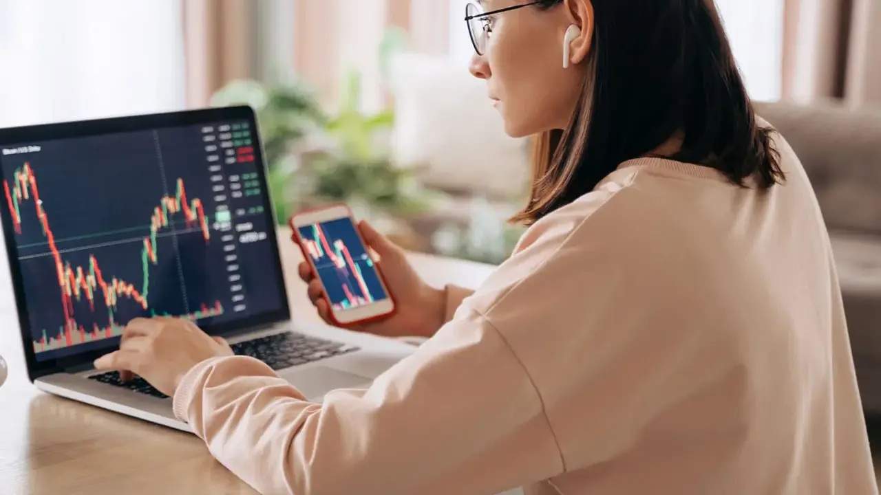 Donna analizza grafici finanziari su laptop e smartphone, cercando il significato delle speculazioni.