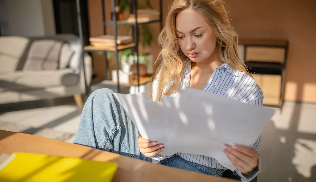 Donna bionda studia documenti, cercando di capire gli oneri accessori. Luce solare filtra attraverso le persiane.