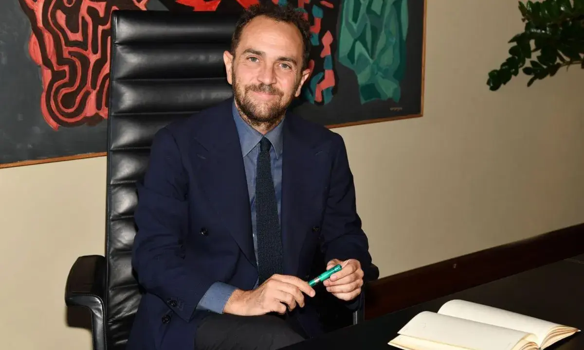 Uomo in giacca blu seduto alla scrivania, con un libro aperto e una penna verde. Un quadro astratto alle sue spalle, che ci fa riflettere su cosa sono le istituzioni.