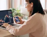 Donna analizza grafici finanziari su laptop e smartphone, cercando il significato delle speculazioni.