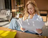 Donna bionda studia documenti, cercando di capire gli oneri accessori. Luce solare filtra attraverso le persiane.
