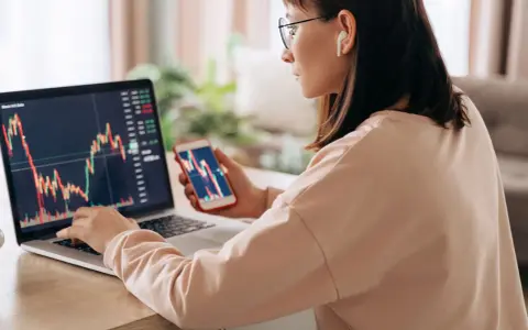 Donna analizza grafici finanziari su laptop e smartphone, cercando il significato delle speculazioni.