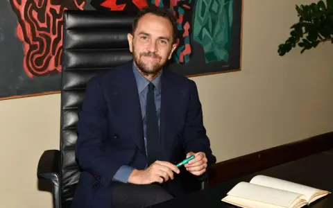 Uomo in giacca blu seduto alla scrivania, con un libro aperto e una penna verde. Un quadro astratto alle sue spalle, che ci fa riflettere su cosa sono le istituzioni.