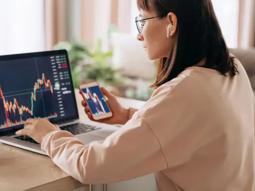 Donna analizza grafici finanziari su laptop e smartphone, cercando il significato delle speculazioni.