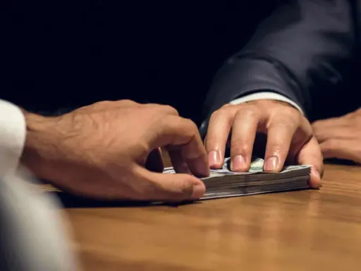 Mani che si scambiano denaro su un tavolo. Un uomo in giacca e cravatta porge una mazzetta di banconote, l'altro le afferra. Il significato di estorsione è chiaro.