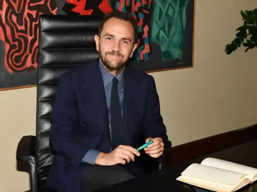 Uomo in giacca blu seduto alla scrivania, con un libro aperto e una penna verde. Un quadro astratto alle sue spalle, che ci fa riflettere su cosa sono le istituzioni.