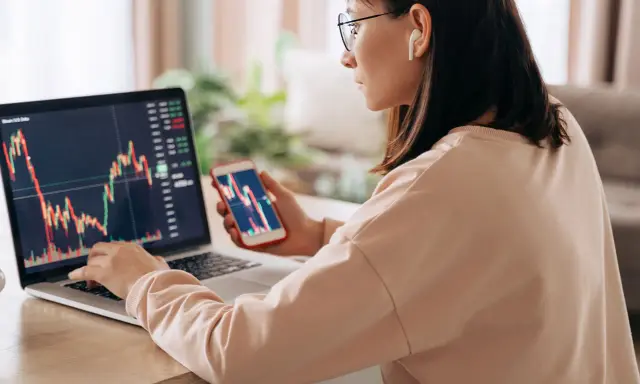 Donna analizza grafici finanziari su laptop e smartphone, cercando il significato delle speculazioni.