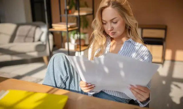 Donna bionda studia documenti, cercando di capire gli oneri accessori. Luce solare filtra attraverso le persiane.