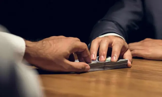 Mani che si scambiano denaro su un tavolo. Un uomo in giacca e cravatta porge una mazzetta di banconote, l'altro le afferra. Il significato di estorsione è chiaro.