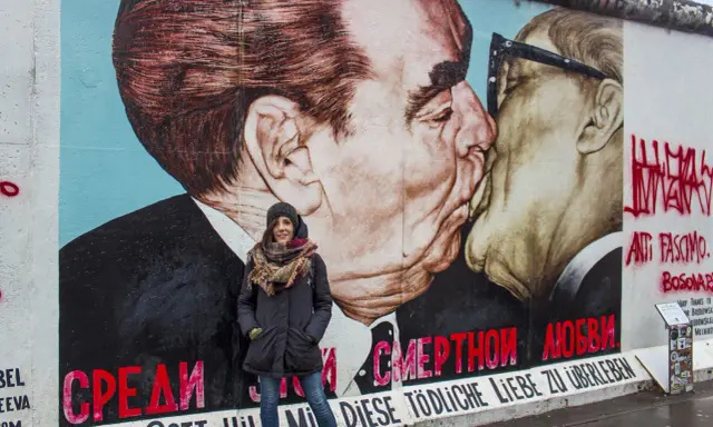 Donna sorride davanti al murale "Il bacio" sull'East Side Gallery, simbolo delle differenze tra Berlino Est e Ovest.