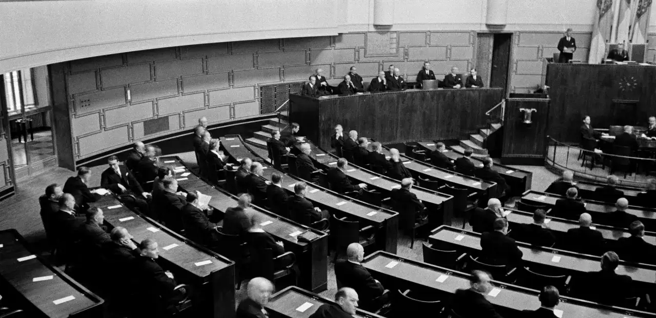 Uomini in abiti scuri siedono in un'aula, rappresentando un dibattito tra liberali e democratici.