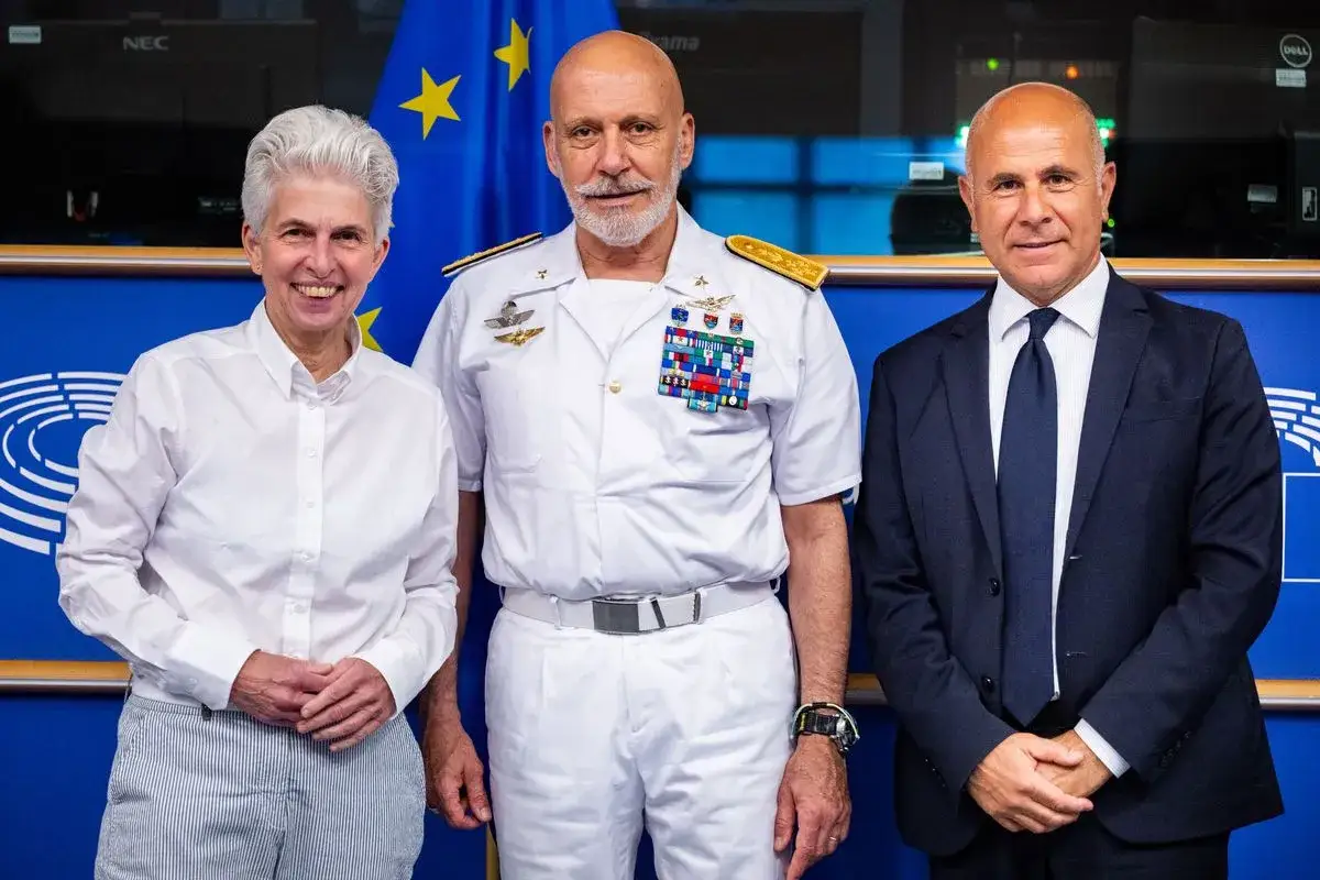 Tre persone in piedi davanti alla bandiera europea. Al centro un ammiraglio con molte medaglie, simbolo di una forte difesa europea.